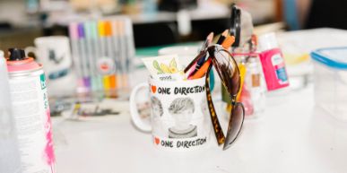 Nahaufnahme einer Tischfläche mit verschiedenen Schreib- und Malutensilien. In der Mitte eine Tasse mit einer Sonnenbrille.