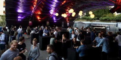 Ein grosses schwarzes Zelt spannt sich über einen Platz. Es stehen Menschen verteilt an Stehtischen darunter und sprechen miteinander.