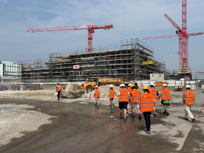 Baustelle mit halbfertigem Rohbau im Hintergrund, im Vordergrund Projektleiter mit einer Gruppe Studierender - alle mit Warnwesten und Helmen