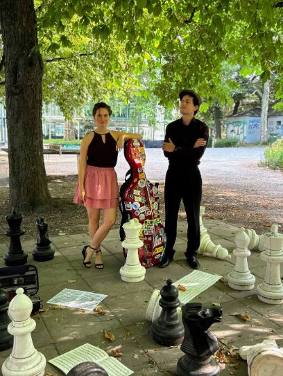 Zwei Menschen mit einem Cellokoffer stehen auf einem Schachfeld unter einem Baum. 