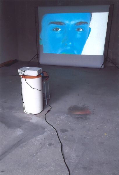 Aufnahme des Ortes der Installation: verfleckter Betonboden, weisser (Eimer) mit Projektor und zuhinterst im Bild die Leinwand mit einem blauen Jungengesicht.