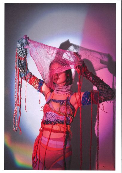 Eine junge Frau in rötlichem Licht mit diversen Strumpfhosen und Unterhosen und gestrickten Ärmeln und Büstiers, einem Netzstoff vor dem Gesicht haltend. an den hochgehaltenden Händen und Armen hängen rote Fäden herunter.