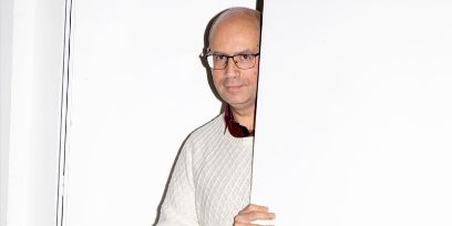 Weisses Bild. Der Mann in weissem Pullover schaut hinter einer Türe oder einer Wand hervor. Aufnahme von der Brust aufwärts. Er trägt eine Glaze und eine Brille.