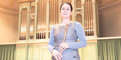 La femme d'âge moyen aux cheveux bruns se tient devant un orgue et tient sa flûte traversière près de son corps. Elle porte des vêtements clairs dans les tons bleu-gris.