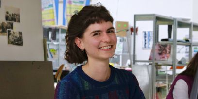 Die junge, braunhaarige Frau trägt einen Pulli in Blautönen und blickt leicht seitlich zur Kamera. Sie sitzt in einem Atelier/Büro.