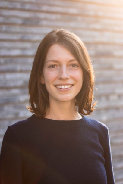 Portrait der braunhaarigen Frau. Sie lächelt in die Kamera, trägt die schulterlangen Haare offen und einen blauen Pullover. Die Sonne scheint von rechts oben.