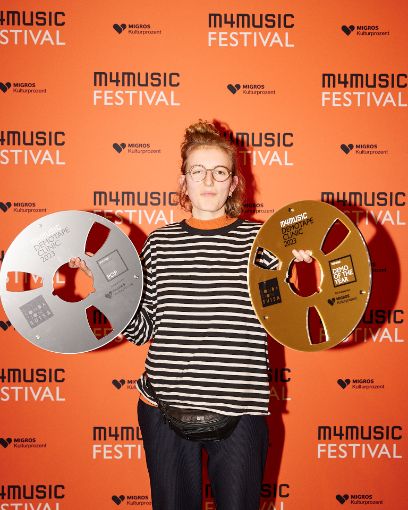 Vor orangenem Hintergrund auf dem steht m4music Festival steht die junge dunkelblonde Frau mit halboffenem Haar, Brille und den beiden Platten/Gewinnen in der Hand