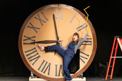 Eine Frau im Blaumann steht auf der Bühne vor einer riesigen Uhr und hält die Mitte der Zeiger fest und spielt mit ihrem Körper Uhrzeiger (rechtes Bein in gegen 9 Uhr, Meter in der linken Hand des in die Höhe gestreckten Arms).