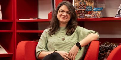 La jeune femme est assise dans un fauteuil rouge devant une bibliothèque rouge. Elle porte un pull vert clair, des lunettes et ses cheveux bruns sont détachés. Elle a le bras gauche posé sur l'accoudoir. Elle sourit à la caméra, la bouche fermée.