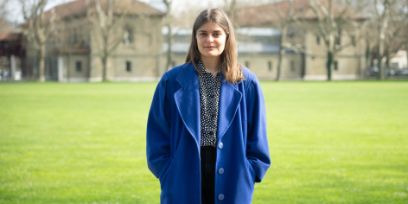 Une femme avec un manteau bleu et des cheveux bruns se tient devant une prairie verte et regarde la caméra. En arrière-plan, on voit des arbres sans feuilles et des bâtiments.