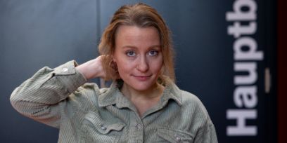 Die dunkelblonde Frau trägt ein Hemd, hält ihre Haare mit der rechten Hand im Nacken fest und blickt freundlich in die Kamera. Im Hintergrund ist eine Türe und der senkrechte weisse Schriftzug «Hauptb» zu sehen.