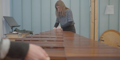 Une femme se tient à l'extrémité d'un marimba et travaille dessus. Au premier plan à gauche, on voit vaguement une main.