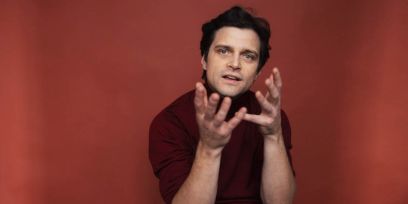 Ein Mann mit braunen Haaren schaut in die Kamera und gestikuliert mit beiden Händen.