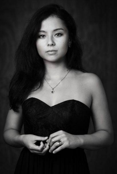 Eine Frau schaut direkt in die Kamera. Sie hat dunkle Haare und ein schwarzes Kleid an, sie trägt eine Halskette mit Anhänger. Das Bild ist in schwarz-weiss.