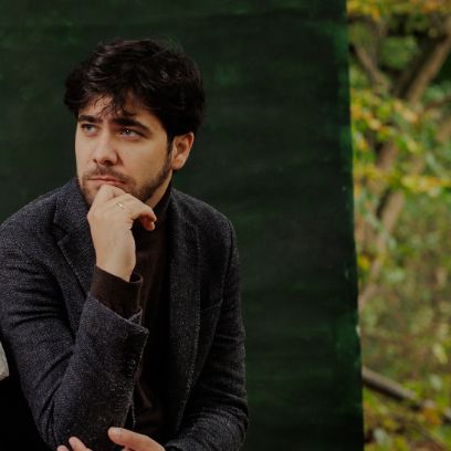 Ein Mann hält sich das Kinn und schaut schräg an der Kamera vorbei. Er hat dunkle Haare und einen Bart. Er trägt dunkle Kleidung.