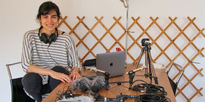 Die junge Frau ist in der Hocke auf einem Stuhl. Ihren linken Arm hat sie auf einem Holztisch, auf dem diverse technische Geräte stehen: Laptop, Stativ, verschiedene Mikrofone usw.