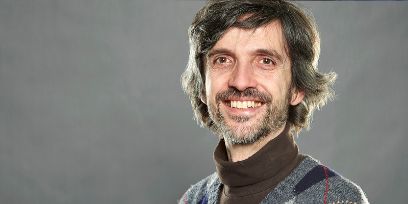 Porträtaufnahme von Claudio Bacciagaluppi, braun-graues Bart- und Kopfhaar. Er lächelt mit offenem Mund in die Kamera. Grauer Studio-Hintergrund.