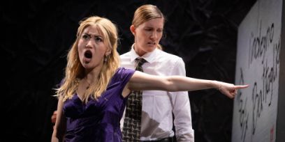 Zwei Personen stehen auf der Bühne. Die vordere Person hat den Mund offen und schreit. Dabei zeigt sie nach links. Die zweite Person steht hinten und schaut auf den Boden.