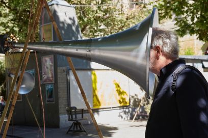 Ein Schalltrichter unter Bäumen, im Hintergrund eine Plakatwand. Ein Mann hält den Kopf halb in den Schalltrichter.