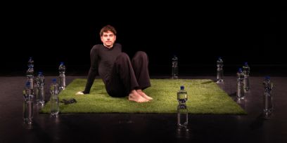 Le jeune homme porte des vêtements noirs moulants et est assis, les jambes repliées et les pieds tendus, sur un carré de faux gazon dans une pièce sombre. Il s'appuie sur ses mains. Autour du gazon sont disposées quelques bouteilles en plastique sans étiquette, munies d'un bouchon bleu.