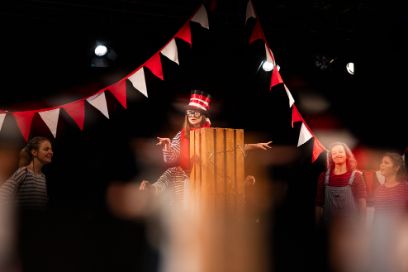 Eine verkleidete Frau steh auf der Bühne. Vor ihr sieht man eine Holzkiste.