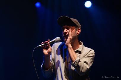 Ein Mann hat ein Mikrofon in der Hand und singt. Er trägt eine Mütze und ein graues Hemd.