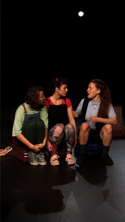 Drei junge Frauen sitzen auf einer Kiste auf einer Bühne. Die Frau links singt, die beiden anderen Frauen schauen sie an.
