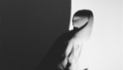 Unscharfes Schwarzweiss-Bild einer in eine Kapuzenjacke gehüllte Person, die sich halb im Schatten befindet. Das Gesicht ist abgewendet.