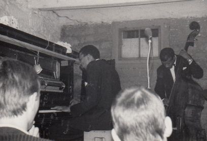 A black pianist on the left and a black bassist on the right play music in a basement in front of a white audience.