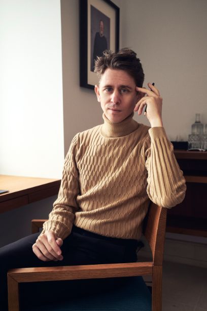 Der jung Mann sitzt in einem Stuhl am Fenster und hält die rechte Hand am Kopf. Er trägt einen beigen Pullover und eine dunkle Hose.