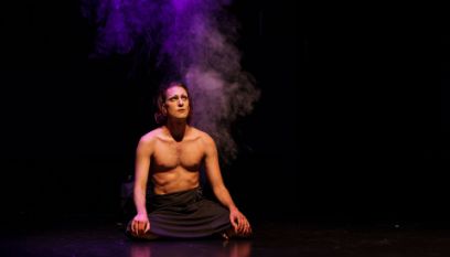 Geschminkte Person mit nacktem muskulösem Oberkörper sitzt auf der Bühne und schaut nach oben, dahinter violetter Rauch
