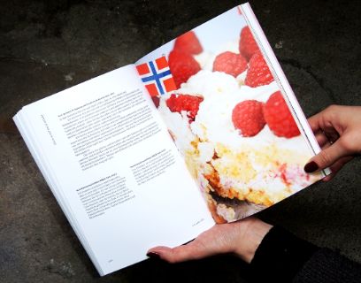 Zwei Hände halten ein aufgeschlagenes Buch vor dunklem Hintergrund. Auf der linken Seite des Buches ist Text zu sehen, auf der rechten ein Foto.