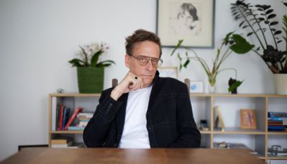 Mann mit Brille und Anzug sitzt vor pflanzenbestücktem Regal an einem Holztisch und schaut, auf seine beringte Hand bestützt, in die Kamera