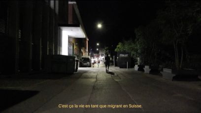 Ein nächtliche Strasse mit beleuchteten Hauseingängen. In der Mitte des Bildes ist von hinten die Silhouette einer Person zu sehen. In der Untertitelung steht: C'est ça la vie en tant que migrant en Suisse.