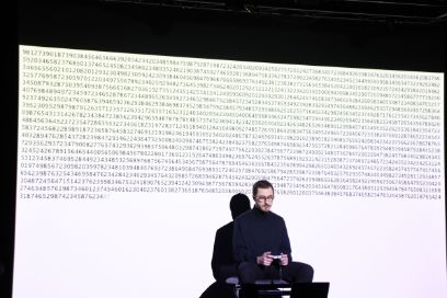 A young man dressed in black sits with a game controller in his hand in front of a series of numbers projected onto a screen.