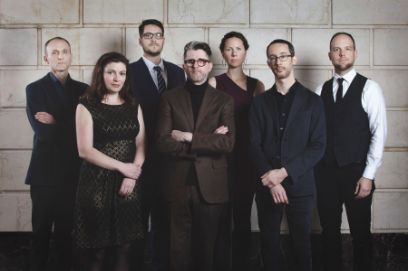 Sieben dunkel gekleidete Frauen und Männer mittleren Alters stehen vor einer Mauer mit grossen hellgrauen Quadern.