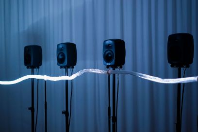 In einer Reihe auf Stativen aufgestellte Lautsprecher, durch eine LED-Lichterkette verbunden, stehen blau ausgeleuchtet vor einem Vorhang.