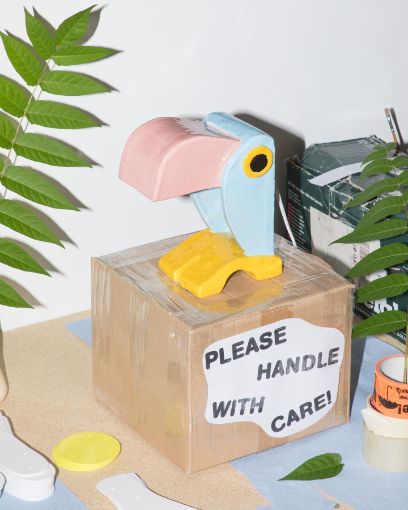 Foto, bunter Keramikvogel auf Kartonkiste Aufschrift "please handle withe care"