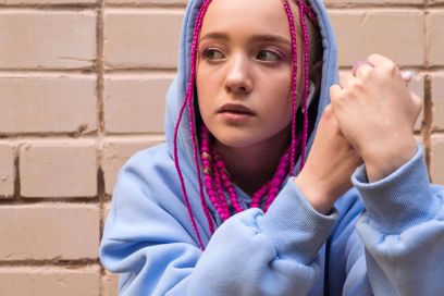 Ein junges Mädchen mit blauem Pullover und violetten Rastazöpfen sitzt nachdenklich an einer Wand. Sie sieht traurig aus.