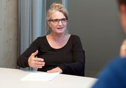 Eine Frau mit Brille und schwarzem Oberteil sitzt an einem Tisch und macht mit der Hand eine erklärende Geste.