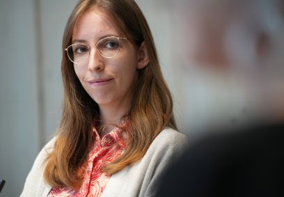 Eine rothaarige Frau mit Brille sitzt und blickt schweigend nach rechts. Sie trägt eine orange Bluse.
