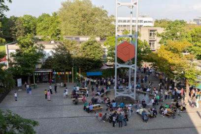 Foto des Platzes rund um das Glockenspiel im Tscharnergut, man sieht Menschen gemeinsam an Festbänken sitzen.