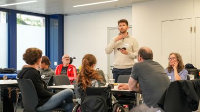 Ein junger Mann erklärt stehend einer Gruppe Sitzender etwas in einem Seminarraum.