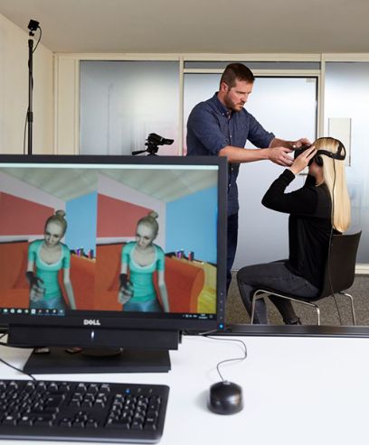Virtual Reality wird als didaktische Methode in der Lehre eingesetzt: eine Studentin zieht mit Hilfe eines Dozenten die VR-Brille an, im Vordergrund ein Bildschirm, der das virtuelle Setting abbildet.