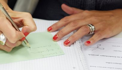 Eine Person mit roten Fingernägeln schreibt etwas auf ein Blatt Papier.