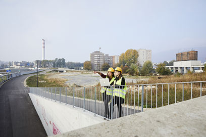 Zwei Lernende mit Helm, Schutzweste und Laptop auf einer Baustelle.