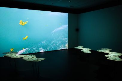 Ein Raum mit einer Leinwand, die das Meer abbildet. Im Vordergrund sind geschwungene Tische sichtbar.