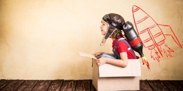 Ein Kind spielt Pilot mit einer kleinen Rakte auf dem Rücken in einem Karton.
